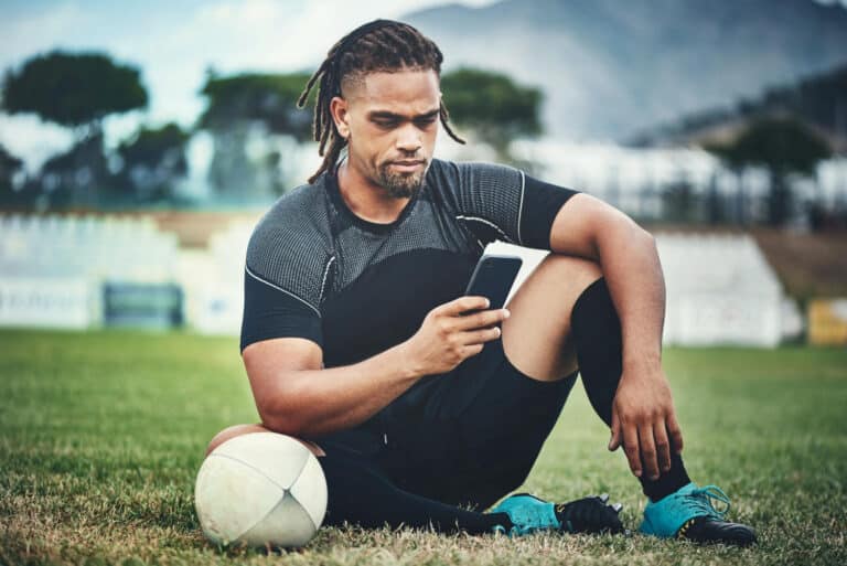 An athletic man with dreadlocks sits on a grassy soccer field, wearing a black and gray athletic shirt and black shorts, looking at his smartphone with a white soccer ball resting by his knee.