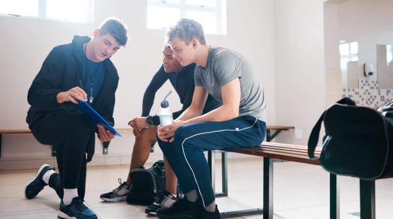Three athletic young men sit on a wooden bench in a bright locker room, with the leftmost kneeling and writing on a blue clipboard while the other two lean in to look at it, one holding a water bottle.
