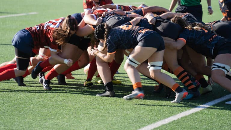 Rugby players in a tight scrum on a green field, interlocked and bent over, wearing red and navy kits against dark blue with orange trim.