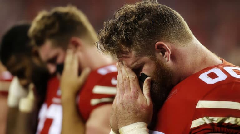 A bearded football player in a red uniform covers his face with his hands on the field.