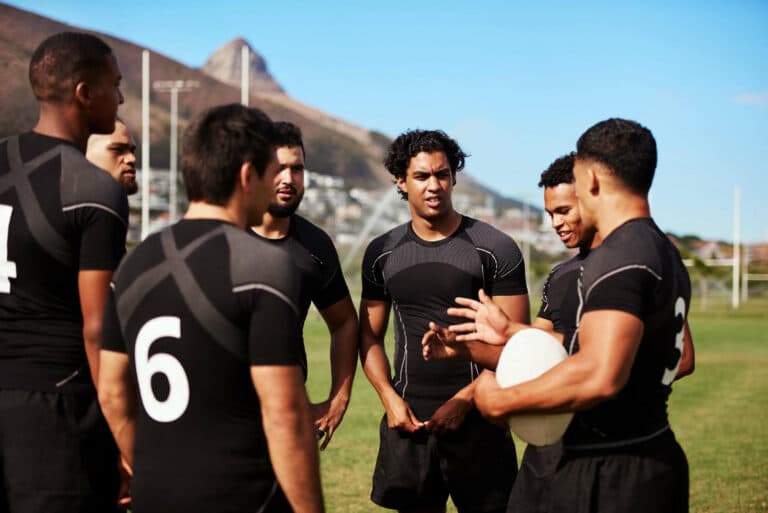 Rugby teammates in black uniforms huddle on a sunny grassy field, one holding a white rugby ball, with mountains and a town in the background.