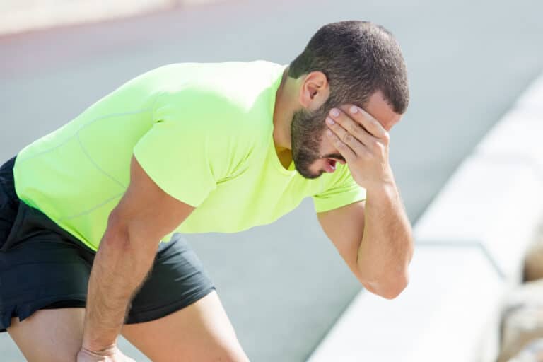 Man outdoors in a neon green shirt and black shorts bends over, head in his hand, looking fatigued.