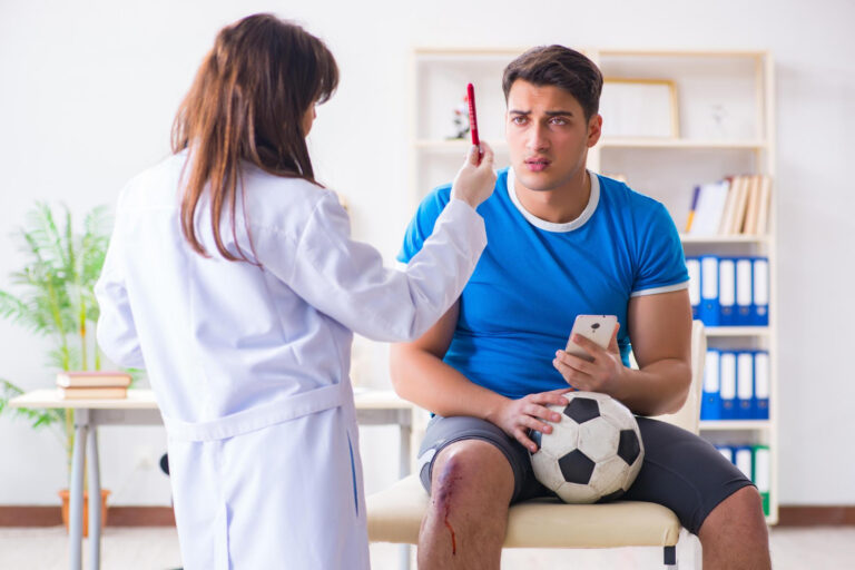A male athlete sits on an examination table holding a soccer ball and a smartphone, while a clinician in a white coat examines his forehead with a red medical tongue depressor. He has a visible leg wound with blood.