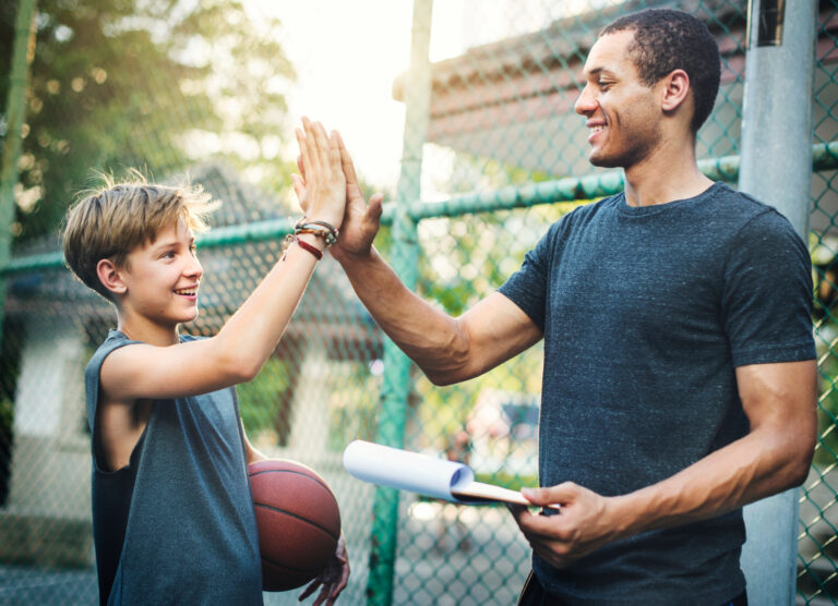 A smiling man and boy give each other a high five on a basketball court, the boy holding a ball and a rolled-up clipboard, as they exchange encouragement.