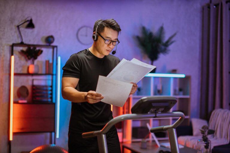Man in a black athletic shirt wearing a headset with a microphone, holding papers while standing at a treadmill in a colorful, modern home gym.